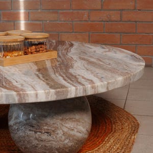 Op de afbeelding: Een ronde marmeren salontafel met een lichtbruin en wit patroonblad en een bolvormige voet. De tafel staat op een geweven jute vloerkleed met oranje accenten. Glazen potten met houten deksels en een houten dienblad staan op de tafel.