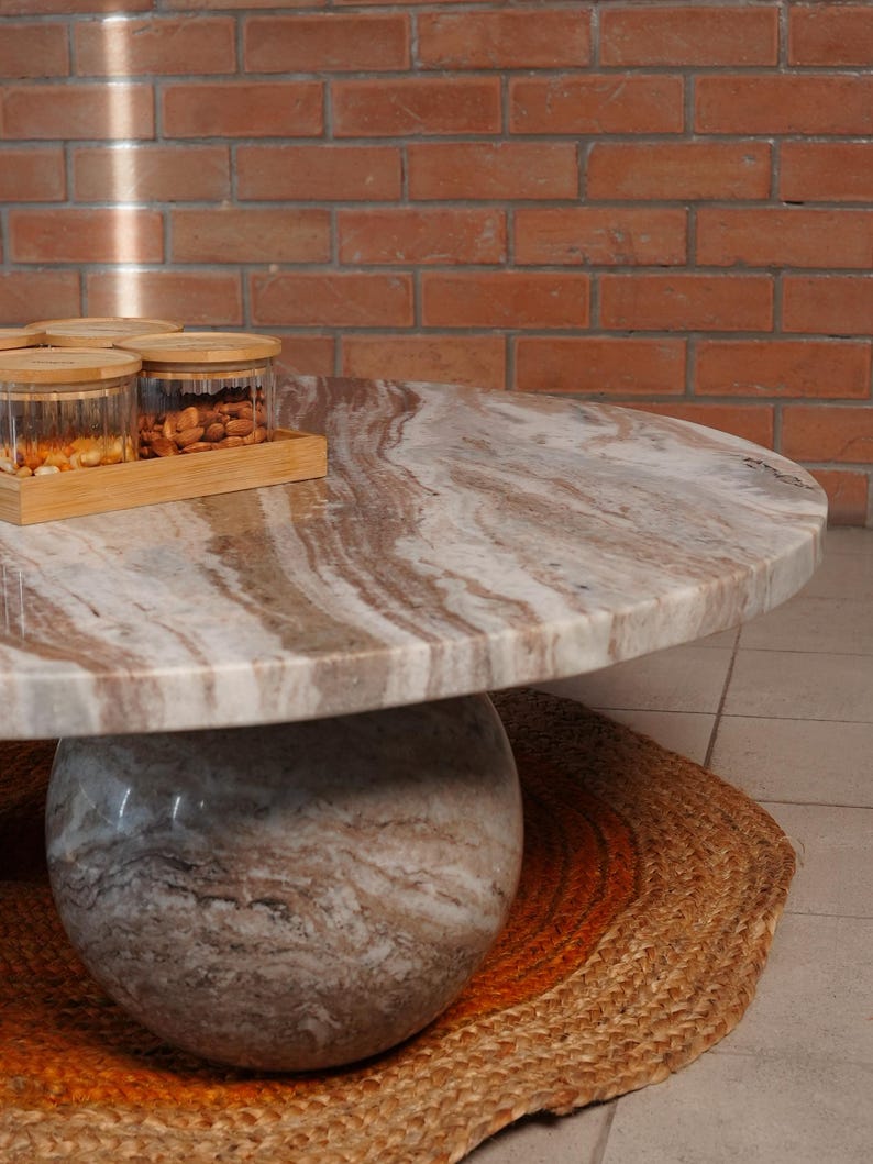Op de afbeelding: Een ronde marmeren salontafel met een lichtbruin en wit patroonblad en een bolvormige voet. De tafel staat op een geweven jute vloerkleed met oranje accenten. Glazen potten met houten deksels en een houten dienblad staan op de tafel.