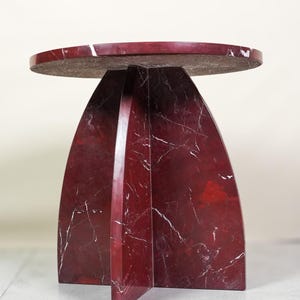 May include: A modern, round side table with a deep red marble finish and white veining. The table has a unique base design with arching supports. The tabletop is approximately 18 inches in diameter.