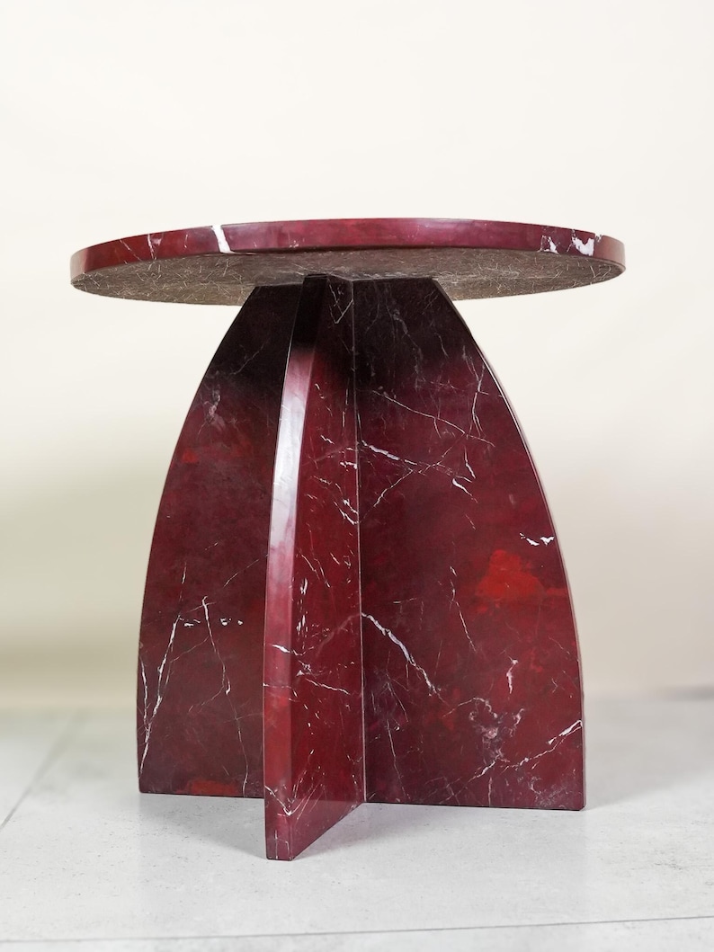 May include: A modern, round side table with a deep red marble finish and white veining. The table has a unique base design with arching supports. The tabletop is approximately 18 inches in diameter.
