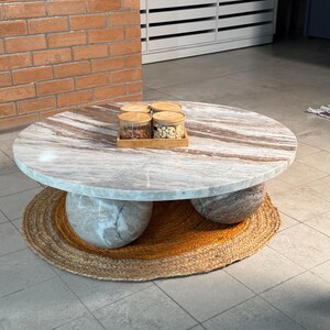 Op de afbeelding: Een ronde marmeren salontafel met een lichtbruin en grijs patroon. De tafel rust op drie bolvormige stenen steunen en een geweven jute vloerkleed. Vier glazen potten met houten deksels worden op een houten dienblad getoond.