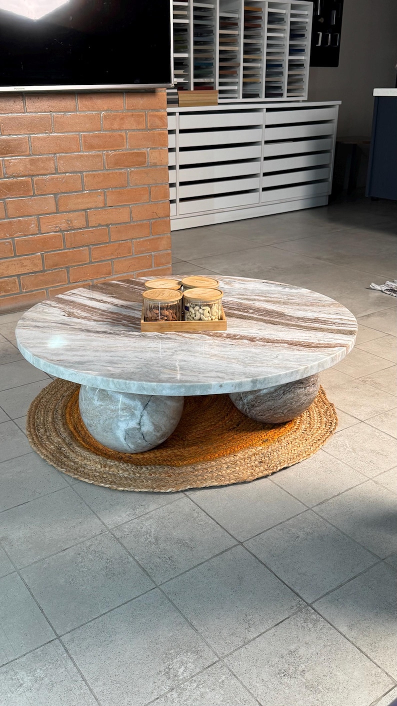 Op de afbeelding: Een ronde marmeren salontafel met een lichtbruin en grijs patroon. De tafel rust op drie bolvormige stenen steunen en een geweven jute vloerkleed. Vier glazen potten met houten deksels worden op een houten dienblad getoond.