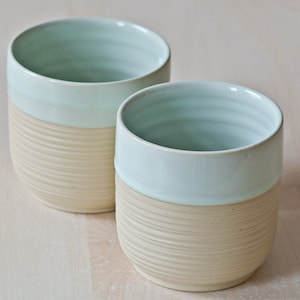 Könnte beinhalten: Zwei Keramikbecher mit hellblauer Glasur und beigefarbenem Boden. Die Becher haben ein strukturiertes, geripptes Design auf der unteren Hälfte.