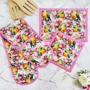 May include: A set of four pink and white kitchen pot holders with a floral print featuring colourful parrots. The pot holders are made of fabric and have a quilted design.