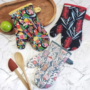 May include: Three oven mitts with different floral patterns. The first mitt has a black background with colourful parrots and flowers. The second mitt has a black background with red flowers and green leaves. The third mitt has a blue background with white flowers and leaves.