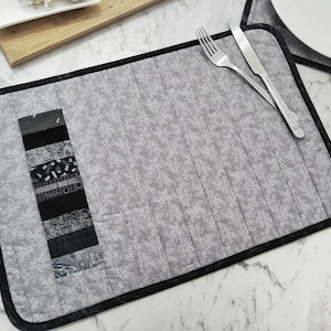 以下が含まれることがあります： 白い花柄と黒い縁取りのある、グレーとブラックのキルティングされたプレースマット。小さなポケットにはカトラリーを入れることができます。