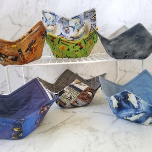 May include: Six fabric bowl cozies in various colours and patterns. The cozies are folded and displayed on a white metal shelf. The patterns include a blue and white fish design, a green and yellow design with tools, a brown and yellow design with construction vehicles, a grey and white design with a vintage newspaper print, a blue and white design with a polar bear print, and a blue and white design with a motorcycle print.