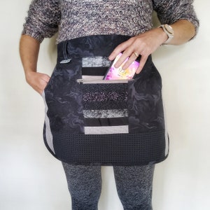 May include: A black and grey quilted apron with a pocket holding a smartphone. The apron features a marbled pattern, a zipped pocket, and a geometric patterned bottom. The apron has a side pocket and a grey trim.