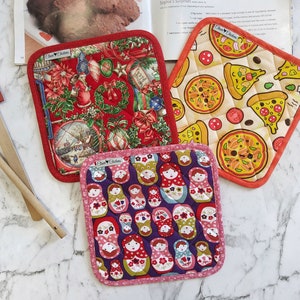 May include: Three red and white quilted pot holders with different designs. One features a Christmas scene, another has a pizza pattern, and the last one has a pattern of Russian nesting dolls.