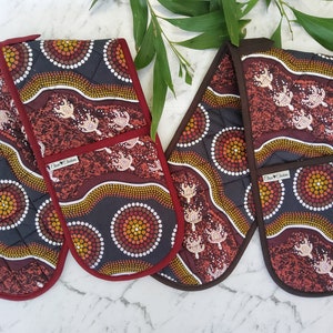 May include: A pair of red and brown oven mitts with an Aboriginal art design featuring circles, waves, and turtles. The mitts are quilted and have a red and brown trim.