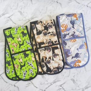 May include: Three oven gloves with dog-themed patterns. One is green with various dog breeds, another features pugs on a black background, and the third has a light blue background with dog silhouettes. Each glove has a black trim.