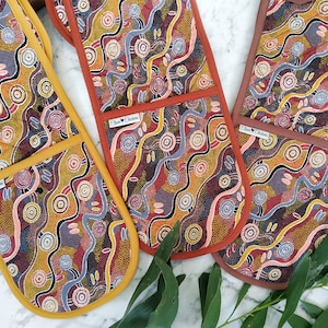 May include: Three oven mitts featuring a colourful Aboriginal art design. The mitts are lined with a contrasting colour fabric. The mitts are on a white marble surface with green leaves.