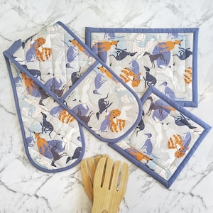 Peut inclure: Un ensemble de cuisine comprenant une manique double, un dessous-de-plat rectangulaire et un dessous-de-plat carré. L'ensemble présente un fond gris clair avec un motif répété de chiens orange, bleus et noirs. Les bords sont bordés de tissu bleu.