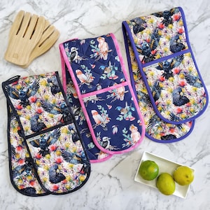 May include: A selection of kitchen textiles with a bird and floral design. The items include double oven gloves with pink and purple trim, and a pair of wooden salad servers. The textiles have a navy blue background.