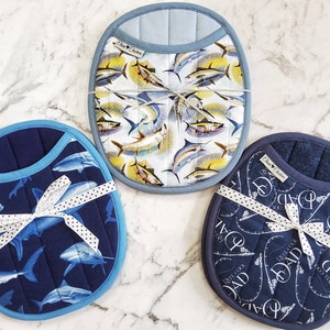 May include: Three blue and white pot holders with a nautical theme. One pot holder features a pattern of fish, another features a pattern of sharks, and the third features a pattern of fishing rods and the word "Dad".