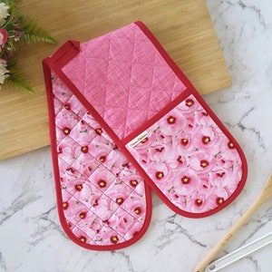 May include: A pair of pink and red oven mitts with a floral print. The mitts are quilted and have a red trim. The text "Oven Mittens" is printed on one of the mitts.