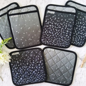 May include: Set of six grey and black quilted pot holders with a geometric pattern. The pot holders have a black trim and a white label that says "Clara Chicken".