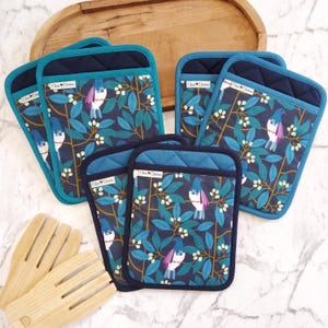 May include: Several quilted pot holders featuring a bird and leaf design in teal, blue, and white. The pot holders have a dark blue quilted top and teal trim. Wooden salad servers are also present.