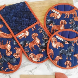May include: A navy blue oven glove and two round pot stands with a dachshund dog pattern. The items have a rust-coloured trim. The oven glove is double-sided. The pot stands are round and feature the same dog pattern.