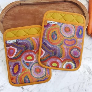 以下が含まれることがあります： カラフルな抽象デザインの長方形のオーブンミトン2つ。ミトンは、マスタードイエローのキルティングトップと、オレンジ、紫、緑の色合いの同心円の鮮やかなパターンが特徴です。ミトンは木製のトレイに置かれています。