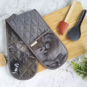 May include: A pair of grey quilted oven mitts with a brown and white animal print. The mitts are on a wooden chopping board with a red spatula and a black spoon.