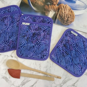 May include: Three quilted pot holders and a square hot pad with a purple batik pattern of white dots on a blue background. The edges are trimmed with a solid purple binding. A wooden spoon and spatula are in the foreground.