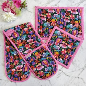 May include: A kitchen set with a floral design on a black backdrop. The set includes an oven mitt, a hot pad, and a square pot holder, all with a pink border. The floral pattern incorporates pink, purple, yellow, and blue. The set is designed for kitchen use.
