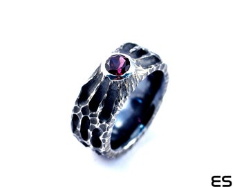Può includere: Un anello d'argento con una finitura testurizzata, ossidata di nero. L'anello presenta una singola pietra preziosa rotonda di colore rosso scuro.