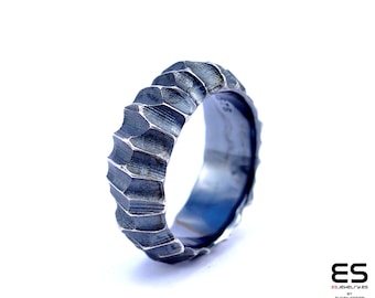 May include: A silver ring with a textured, geometric design. The ring is made of metal and has a dark, oxidised finish.