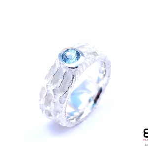 May include: A silver ring with a textured band and a round, light blue gemstone. The ring has a hammered or bark-like texture, and the gemstone is set in a smooth bezel. The ring is displayed against a white background.