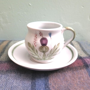 May include: A white ceramic teacup and saucer set. The cup features a hand-painted thistle design with purple, green, and pink accents. The saucer has a green border. The handle of the cup is also green.