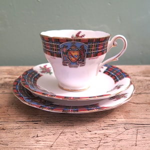Op de afbeelding: Een witte theekop en schotelset met een rood, blauw en groen geruit patroon. De theekop heeft een gouden oor en rand, en een wapen met de woorden "Clan Stewart". De set staat op een houten oppervlak.