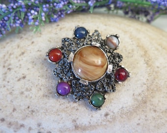 Può includere: Un fermaglio in argento con un nodo celtico, una grande pietra di agata marrone al centro e sei pietre più piccole in rosso, blu, verde e viola.