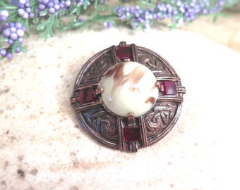 Può includere: Una spilla rotonda color rame con un intricato motivo a intreccio in rilievo. Il centro presenta un cabochon liscio color crema con venature marroni, circondato da quattro piccole pietre sfaccettate quadrate di colore rosso scuro.
