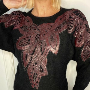May include: Black sweater with a large burgundy lace design on the front. The lace is intricate and detailed, and it covers the entire front of the sweater.