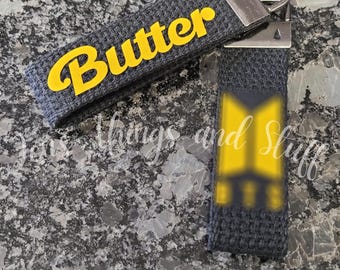 May include: Two dark gray fabric keychains with silver metal hardware. One keychain features the word Butter in bright yellow lettering. The other keychain displays a yellow BTS logo graphic. Both items have a textured, woven appearance.