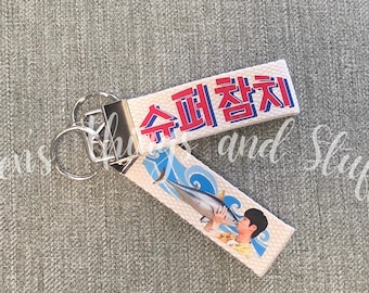 Può includere: Un portachiavi in tessuto con un cinturino bianco che riporta il testo coreano in rosso e blu 슈퍼참치. Il cinturino mostra anche la grafica di una persona che tiene in mano un grosso pesce su uno sfondo di onde blu, fissato a un anello in metallo argentato.