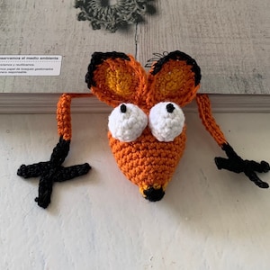 May include: A crocheted orange fox bookmark with black arms and white eyes. The bookmark is hanging off the edge of a book.