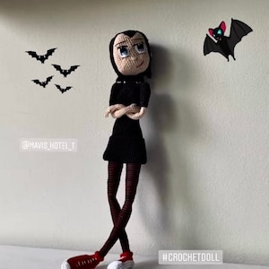 May include: A crocheted doll of Mavis from Hotel Transylvania, wearing a black dress, black and white striped tights, and red shoes. The doll is standing with her arms crossed and looking to the right. There are bats in the background and the text "@MAVIS_HOTEL_T" and "#CROCHETDOLL" are visible.