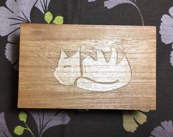 May include: A wooden box with a carved design of a sleeping cat. The cat is white and the box is a light brown colour.