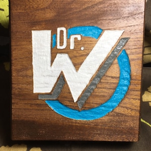 May include: A wooden sign with a carved "Dr. W" logo. The "W" is white with a grey outline and a blue circle around it. The letters "Dr." are white and carved into the wood.