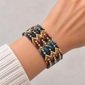 May include: A wide woven beaded bracelet featuring a repeating chevron pattern in shades of deep blue, burgundy, cream, and metallic gold. The intricate design is created with small, uniform glass beads arranged in a dense, multi-row geometric structure.