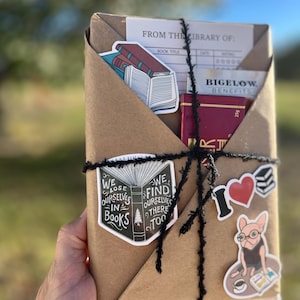 Peut inclure: Un colis en papier kraft envelopp&eacute; de ficelle et d&eacute;cor&eacute; d'autocollants. Les autocollants repr&eacute;sentent un livre avec le texte "We lose ourselves in books. We find ourselves there too." et un chien portant des lunettes et lisant un livre. Le colis comprend &eacute;galement une carte de biblioth&egrave;que avec le texte "From the library of:" et une bo&icirc;te rouge avec le texte "Bigelow Benefits".