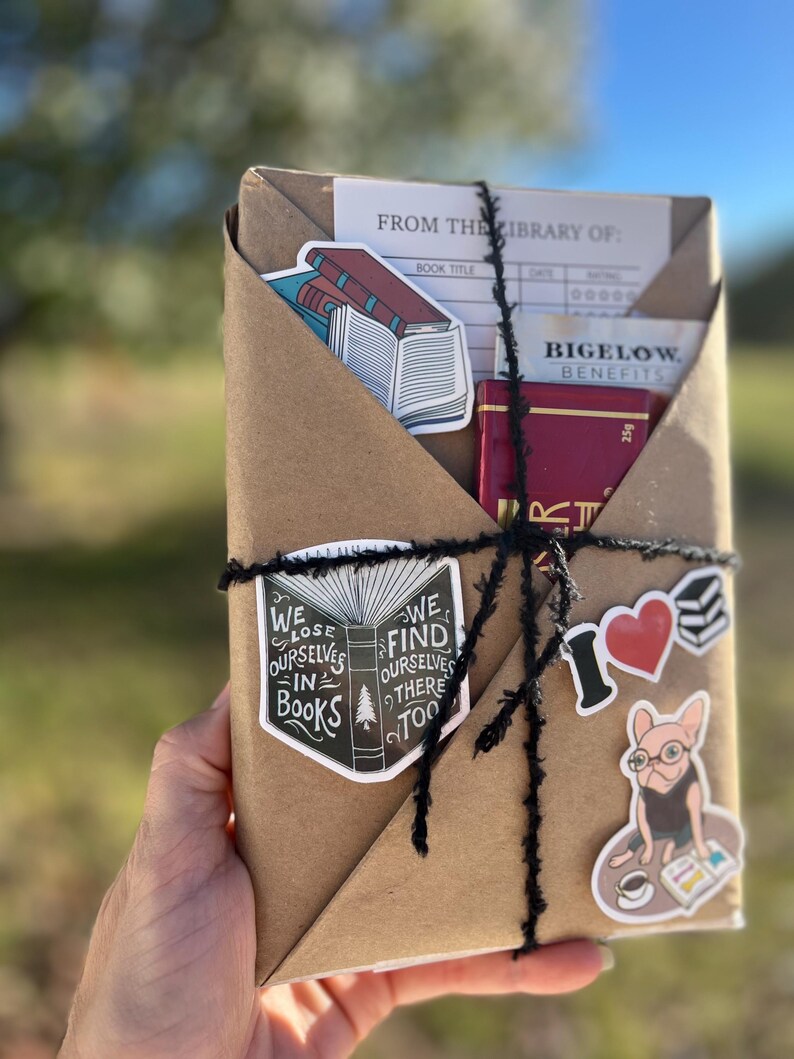 Peut inclure: Un colis en papier kraft envelopp&eacute; de ficelle et d&eacute;cor&eacute; d'autocollants. Les autocollants repr&eacute;sentent un livre avec le texte "We lose ourselves in books. We find ourselves there too." et un chien portant des lunettes et lisant un livre. Le colis comprend &eacute;galement une carte de biblioth&egrave;que avec le texte "From the library of:" et une bo&icirc;te rouge avec le texte "Bigelow Benefits".