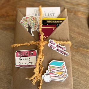 Peut inclure: Un colis en papier kraft envelopp&eacute; de ficelle et d&eacute;cor&eacute; d'autocollants. Les autocollants repr&eacute;sentent un arbre, une tasse de caf&eacute;, une pile de livres et le texte "Hello I'm Reading. Go Away." et "The book was better."
