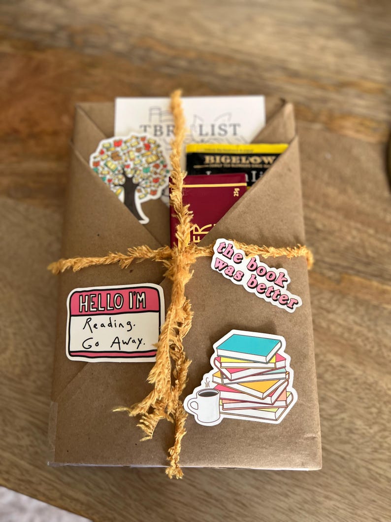 Peut inclure: Un colis en papier kraft envelopp&eacute; de ficelle et d&eacute;cor&eacute; d'autocollants. Les autocollants repr&eacute;sentent un arbre, une tasse de caf&eacute;, une pile de livres et le texte "Hello I'm Reading. Go Away." et "The book was better."