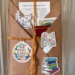 Peut inclure: Un colis en papier brun envelopp&eacute; de ficelle jaune et d&eacute;cor&eacute; de divers autocollants sur le th&egrave;me des livres. Les autocollants comprennent un motif floral, une pile de livres, une main tenant un livre avec le texte "Read More Books" et un cercle avec le texte "Read Books & Grow Plants".