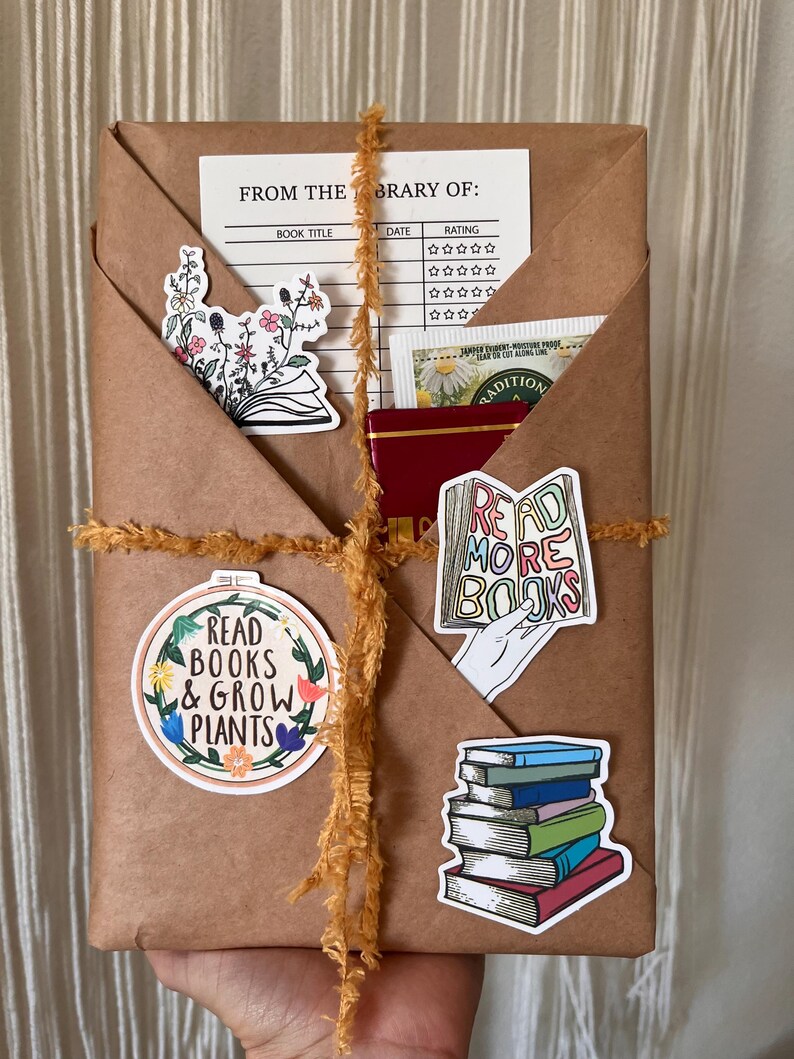 Peut inclure: Un colis en papier brun envelopp&eacute; de ficelle jaune et d&eacute;cor&eacute; de divers autocollants sur le th&egrave;me des livres. Les autocollants comprennent un motif floral, une pile de livres, une main tenant un livre avec le texte "Read More Books" et un cercle avec le texte "Read Books & Grow Plants".