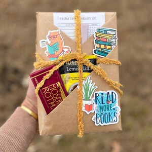 Peut inclure: Un colis en papier kraft envelopp&eacute; de ficelle jaune et d&eacute;cor&eacute; d'autocollants. Les autocollants repr&eacute;sentent un chat lisant un livre, une pile de livres, une plante en pot et un autocollant bleu qui dit "Lisez plus de livres". Le colis contient &eacute;galement une barre de chocolat et un sachet de th&eacute;.