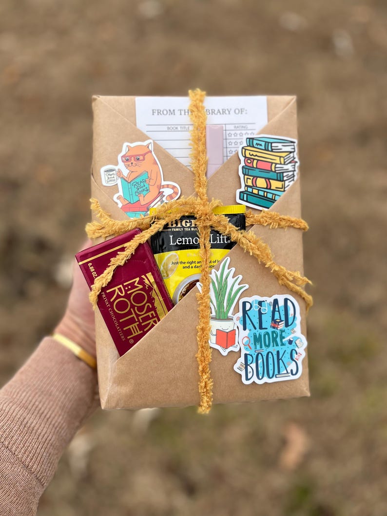 Peut inclure: Un colis en papier kraft envelopp&eacute; de ficelle jaune et d&eacute;cor&eacute; d'autocollants. Les autocollants repr&eacute;sentent un chat lisant un livre, une pile de livres, une plante en pot et un autocollant bleu qui dit "Lisez plus de livres". Le colis contient &eacute;galement une barre de chocolat et un sachet de th&eacute;.